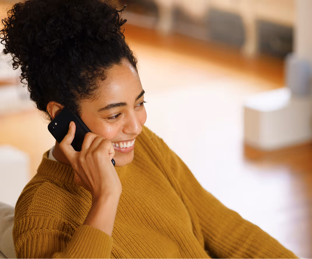 Smiling woman with curly hair wearing a mustard sweater talking on a black smartphone.
