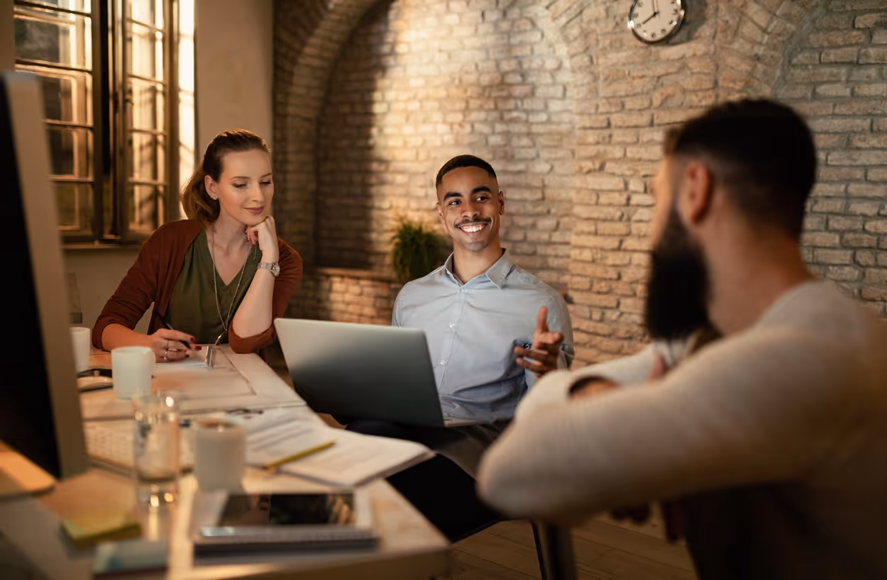 Three young professionals having a discussion around a desk with a laptop and documents in a cozy office.
