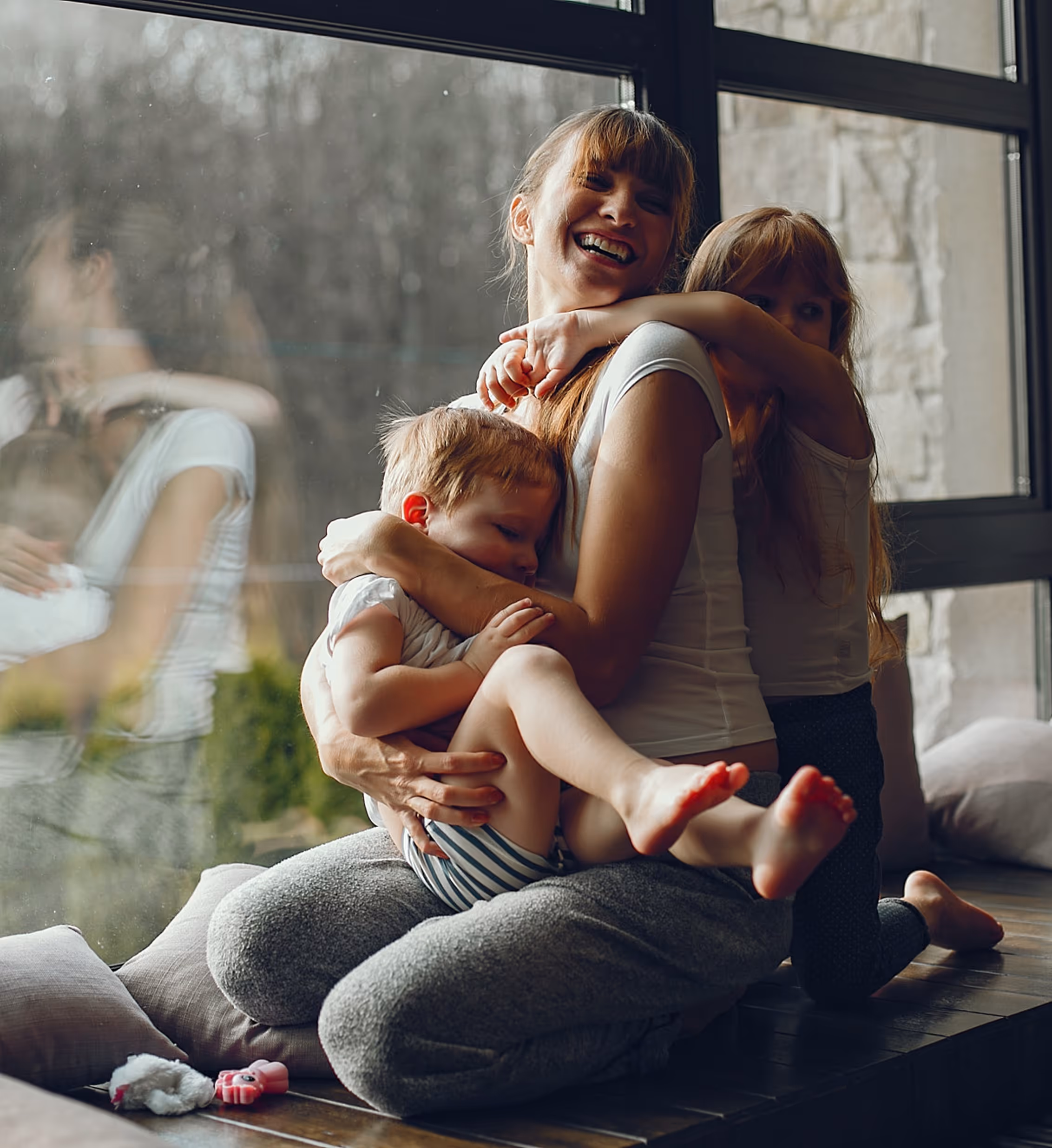 Smiling woman sitting by a window holding a young child while an older child hugs her from behind.