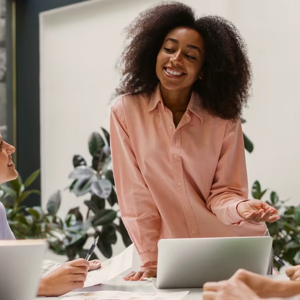 A woman in a pink shirt smiles as she talks with her team around a table with laptops